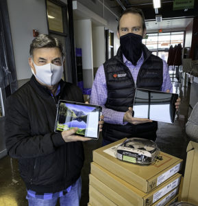 Two men wearing masks holding donated tablets stand behind a table of boxes of headsets.
