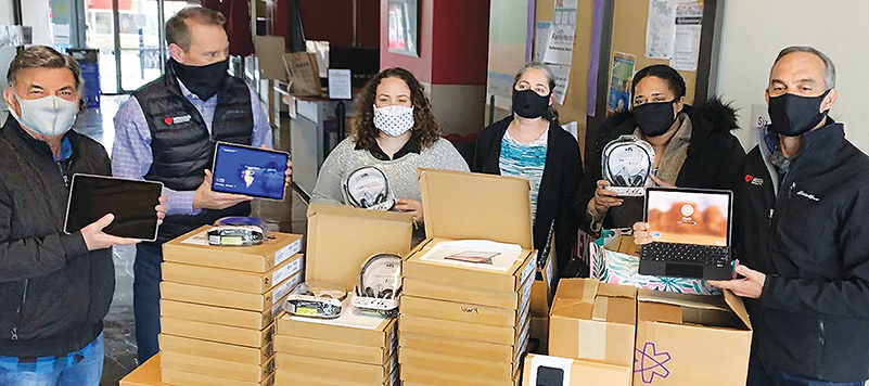 Three men and three women wearing masks pose behind a table of donated tech tools (headsets, tablets, etc.)