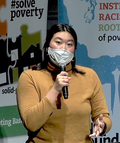 Asian woman in a tan turtleneck sweater and wearing a mask, speaks into a microphone