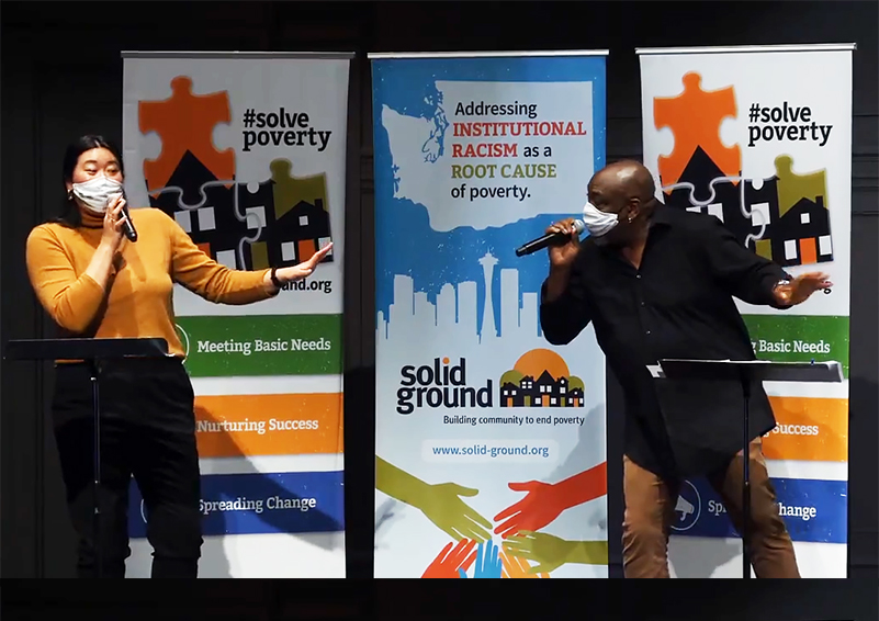 An Asian woman and a Black man, wearing facemasks, sing a socially-distanced duet into microphones