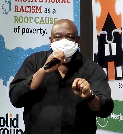 Black man in a black shirt and wearing a mask, speaks into a microphone