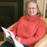 White-haired woman with glasses wearing a pink turtleneck top smiles from a reading chair with a large book open in her lap.