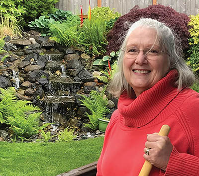 White woman with grey hair and wire-rimmed glasses, wearing a red turtleneck sweater, poses holding a rake in front of her lush, green, landscaped garden and waterfall