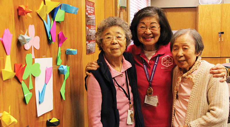 Three senior Japanese woman stand with their arms around each other next to a wall of colorful origami creations
