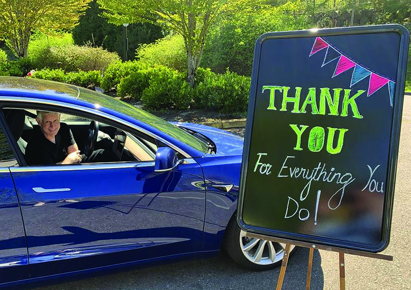 White haired man smiles from his bright blue 4-door sedan next to a colorful chalkboard on an easel reading THANK YOU for everything you do!