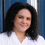 Headshot of a woman with Black hair wearing a white blouse in front of a teal building
