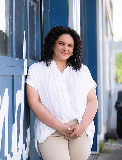 Black woman with curly hair wearing a white blouse and khaki pants stands in front of a blue wall.