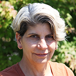 Smiling headshot of a woman with short grey hair wearing an orange top, in front of a green bush