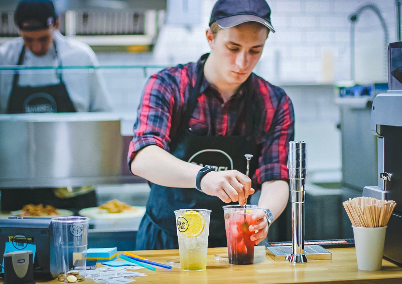 A young man in a plaid shirt stirs a drink while standing behind the counter of a restaurant