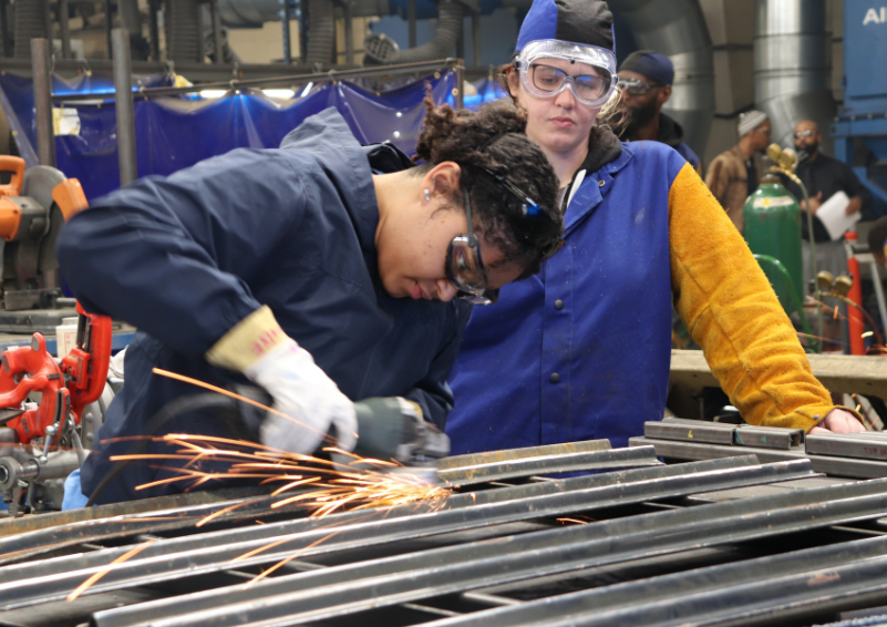 A picture of one woman watching another working in a machine shop as sparks fly.