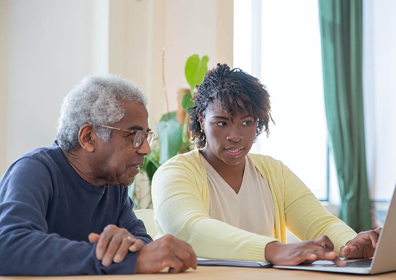 An older man and young woman look at a laptop on a table.