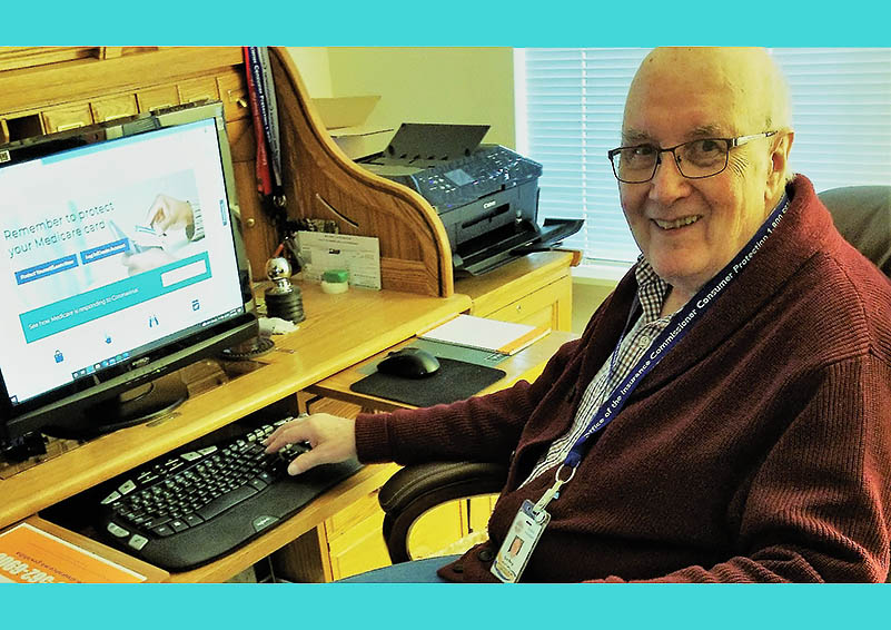 An elderly white man with glasses and a cardigan sits at a computer with Medicare information showing on the screen
