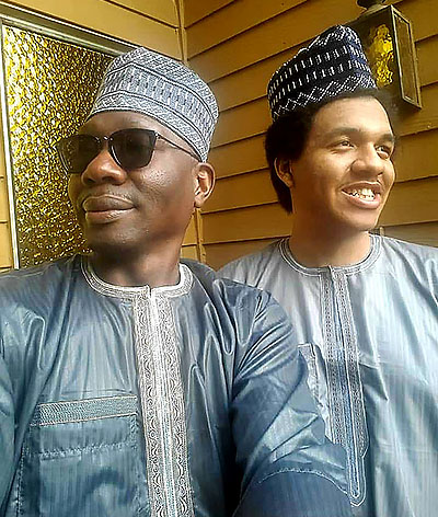 A father and son in light blue Nigerian traditional regalia in front of a yellow wall.