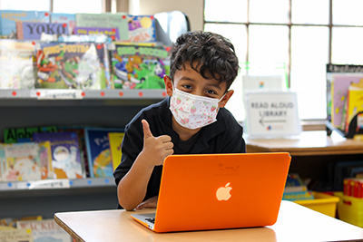 A young boy gives a "thumbs up" from behind a laptop.