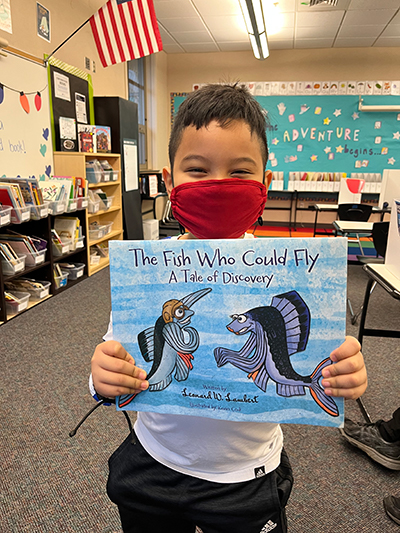 A young boy holding a hand-illustrated book titled: "The Fish Who Could Fly: A Tale of Discovery."