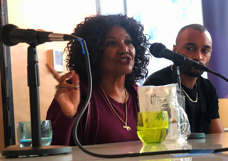 A woman at a table talks into a microphone while a man next to her listens.
