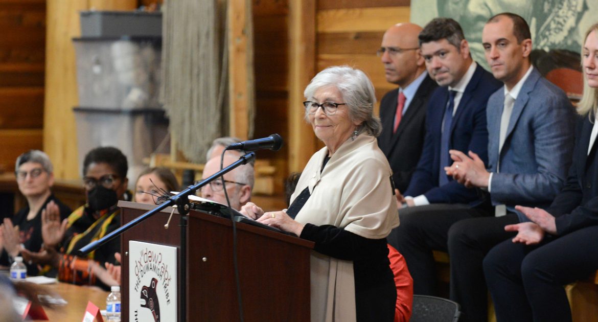 An older woman with white hair and wearing a white shawl, stands at a lectern flanked by men in suits and others off behind.