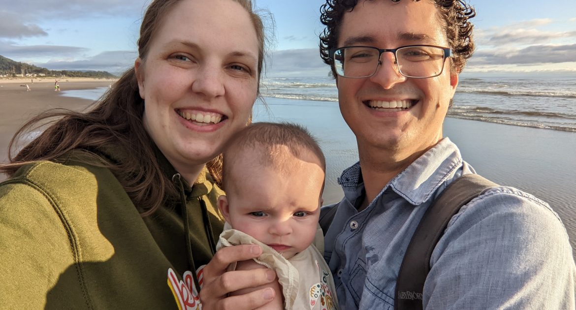 A woman in an olive-green hoodie and a man in a blue collared shirt hold a baby between them as waves lapping the sand behind them.