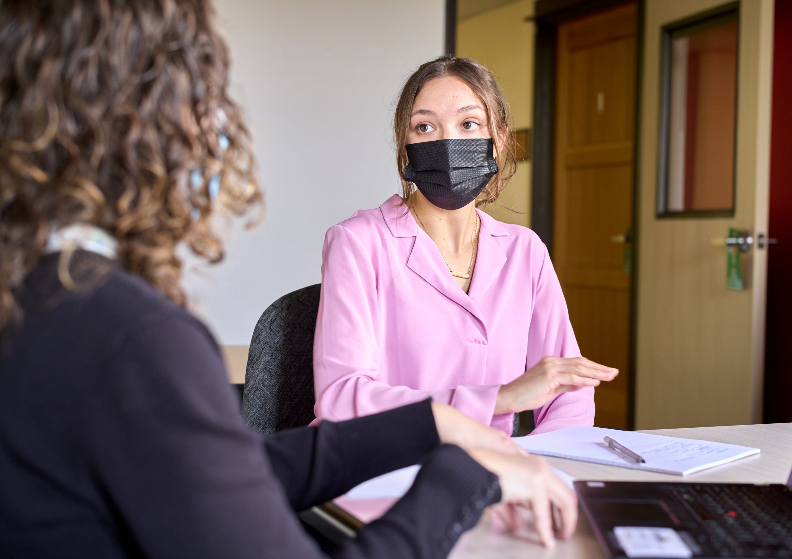 A woman with curly hair and black hair, her back turned to the camera, gestures while taking with a woman in a pink blouse and black mask.