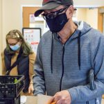 A man wearing a gray hoodie and black mask prepares to fill a paper sack with vegetables.