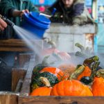 A trough of orange and green squash are sprayed with water from a hose.