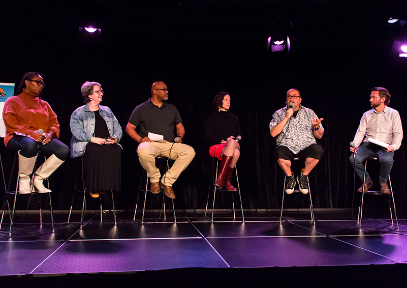 Six people sit on stools on a stage. Five of them are turn to look at the one who is speaking into a microphone.