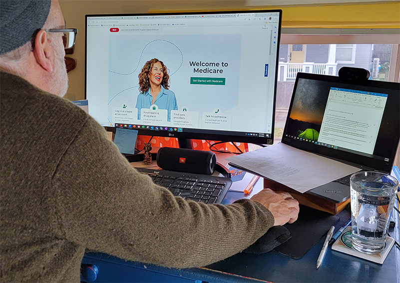 A man in hat, glasses, and brown sweater looks at a computer screen that is open to a webpage that reads "Welcome to Medicare."