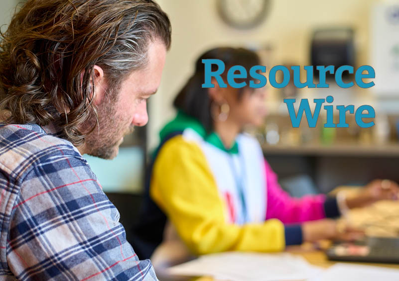 A man with long hair and a blue patterned flannel looks down at something out of shoot. A person can be seen out of focus in the background and the words "Resource Wire" appear besides him.