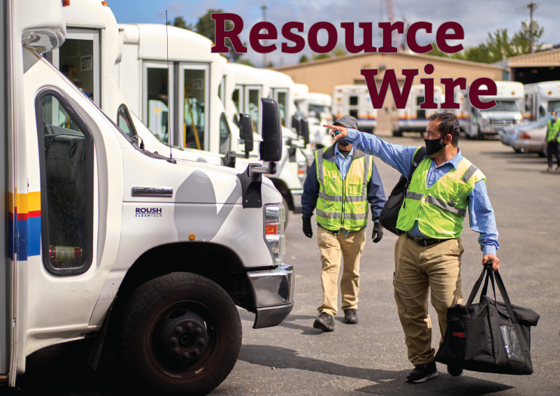 Two men in neon green vests and khaki pants walk down a row of white shuttle buses. The words "Resource Wire" appears above them.