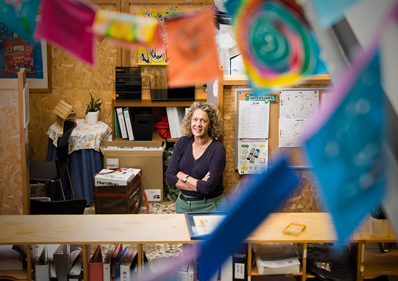 A woman with wavy hair stands with her arms folded in the middle of a cluttered office.