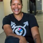 A smiling woman in a trans rights t shirts, showing a combination of the male and female symbols with a fist in the center.