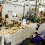 A woman standing behind a display of jewelry and other crafts smiles at another woman in a wheel chair.