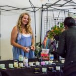 A woman standing behind a display creams and other crafts smiles at a customer looking over her products.