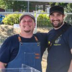 Two men, one in a denim chef's apron, embrace and smile at the camera.
