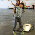 A young boy winds up to throw a bean bag while a woman looks on.