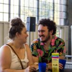 A man with curly hair, a beard, and a Hawaiian shirt lauchs as he talks to a woman in white tank top with her hair in a bun.