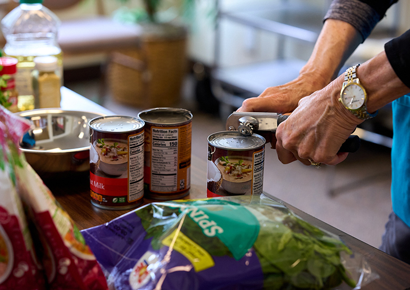 A close up shot of two hands using a can opener to open one of three cans of coconut milk set on a table along with other ingredients.