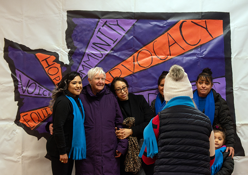 A group of people posing for a picture in front of a large illustration of the outline of Washington state with words like "advocacy," "community," and "voice" radiating out from where Olympia would be on the map.