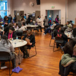 A crowd of people sitting around tables and standing in a conference room.