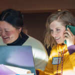 Two woman sit side by side behind laptops, one of them talking on the phone.