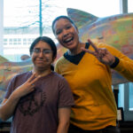 A mother and child flash the peace sign in front of a colorful sculpture of a fish.