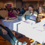 Three people sit at a table -- two of them behind a laptop -- in crowded conference room.