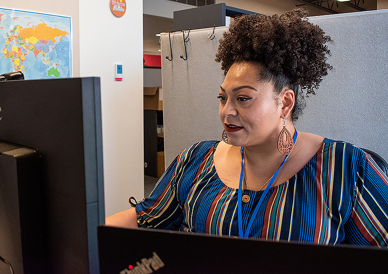 Black woman with curly hair piled on top of her head and wearing a colorful striped top sits in a cubicle in front of a computer monitor.