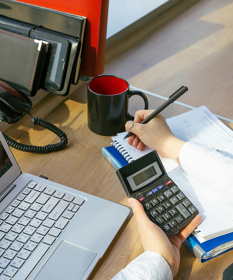 Person with at a laptop holds a calculator in their left hand and writes with a pen on paper with their right hand. There are binders, a phone cord, and a black mug on their table.