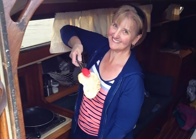 A woman in a blue jacket smiles as she holds up a freshly cooked pita with tongs.