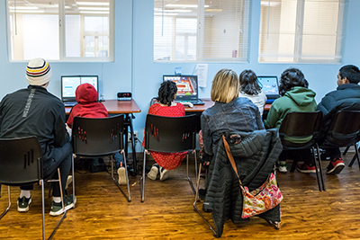 Five kids with their backs to the camera lined up at a desk playing computers games. Two adults sit behind them looking over their shoulders.
