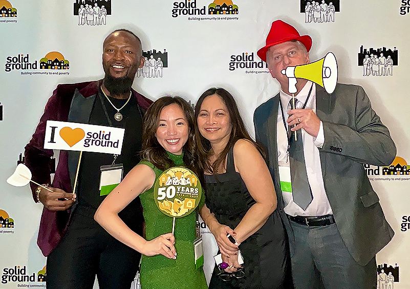 Four people pose in a photobooth, dressed up for a fancy event. One holds a sign that reads I ♥ Solid Ground. Another holds up a circular graphic that reads 50 Years of changing lives. Another wears a bright red hat and holds a megaphone sign up to his mouth.