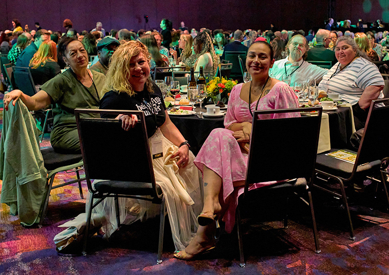 A group of people in fancy dress sitting around a table in a large, crowded ballroom, several them turned in their seats to face the camera.