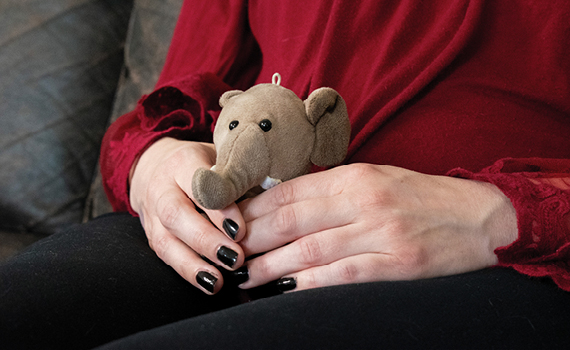 Hands of a woman holding a small stuffed elephant. She wears black nail polish, black pants, and a red blouse.