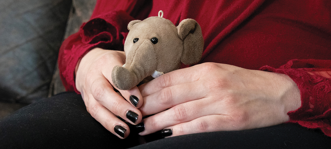 Hands of a woman holding a small stuffed elephant. She wears black nail polish, black pants, and a red blouse.